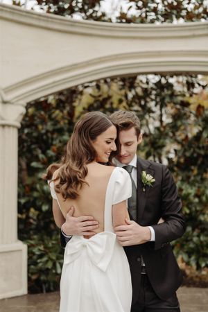 A tender romantic moment at a wedding ceremony with white dress and black suit against an elegant archway and greenery backdrop.