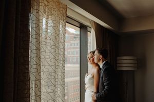 Romantic newlywed couple shares an intimate moment by window with sheer curtains in moody lighting.