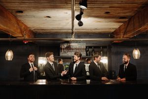 Groomsmen gather at a dark wood bar for celebratory drinks under pendant lighting and exposed wooden beams.
