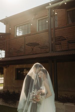 A rustic brick building with outdoor string lights serves as the backdrop for a delicate wedding scene in soft lighting.