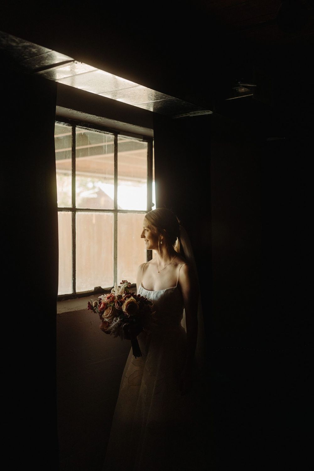 A bride in a white wedding dress stands silhouetted by natural window light while holding a bouquet.