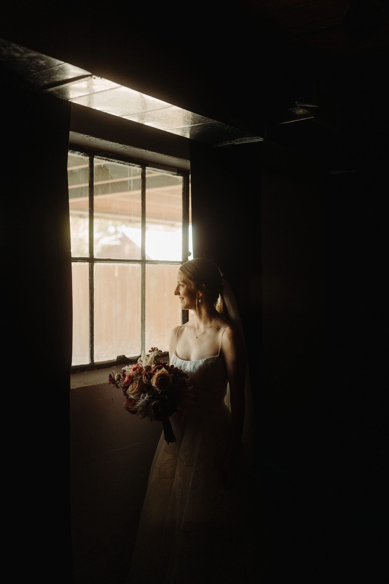 A bride in a white wedding dress stands silhouetted by natural window light while holding a bouquet.