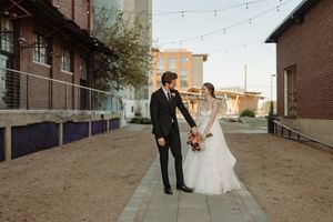 A couple in elegant wedding attire stands together on a city sidewalk with string lights and brick buildings in the background.