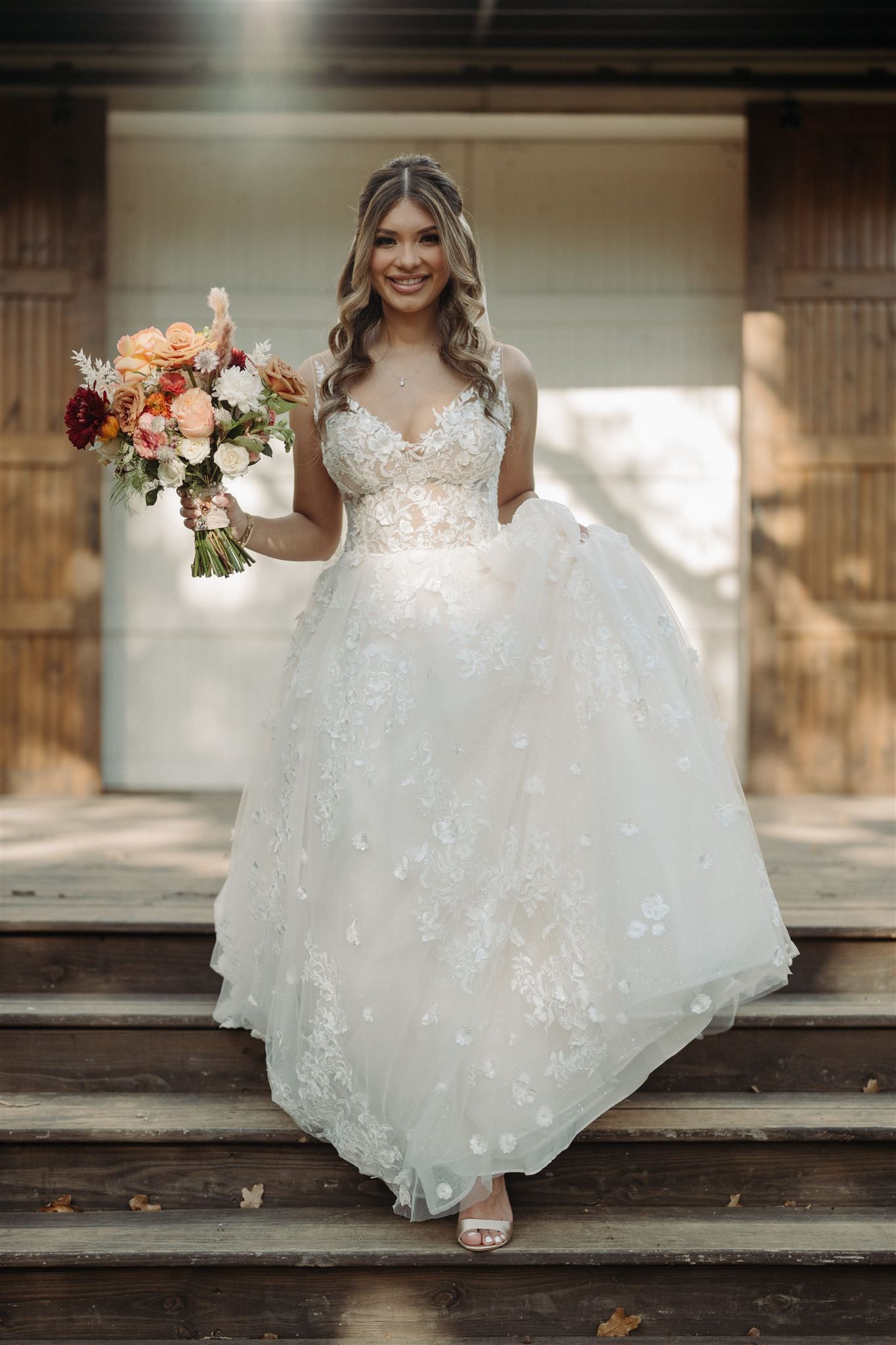 Radiant bride in a sparkling white ballgown stands on rustic wooden steps holding a colorful bouquet.