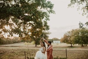 A sunset backlit wedding couple stands side by side in a rustic field with large oak trees in the background.