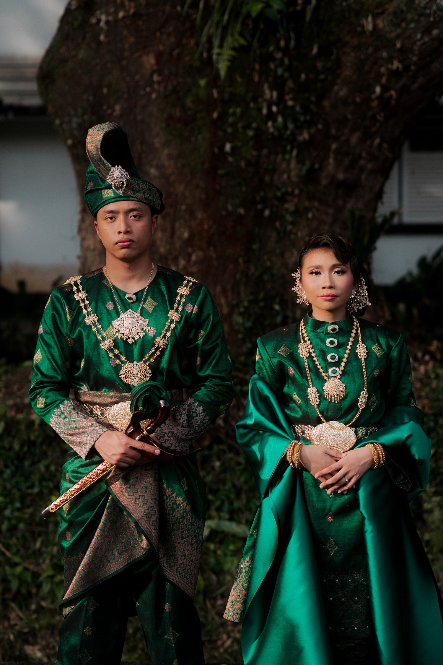 Couple in traditional green Malay royal attire with gold accessories posing outdoors near a large tree.