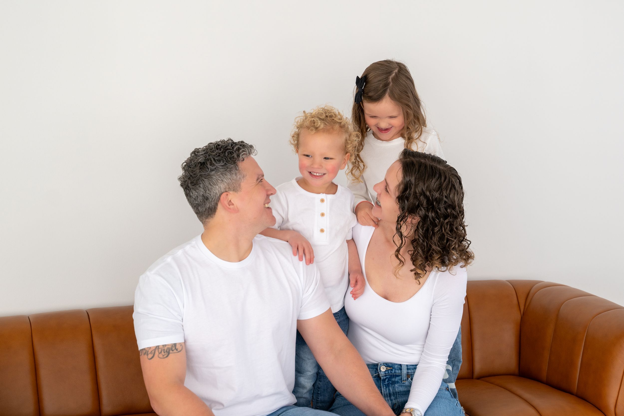 A family wearing white tops sits together on a brown leather couch in a bright room.