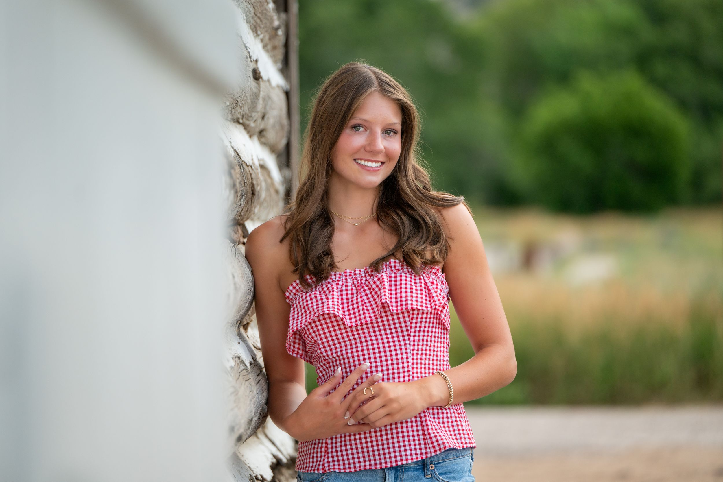 A woman in a red gingham top and jeans poses outdoors by a fence in a natural summer setting.