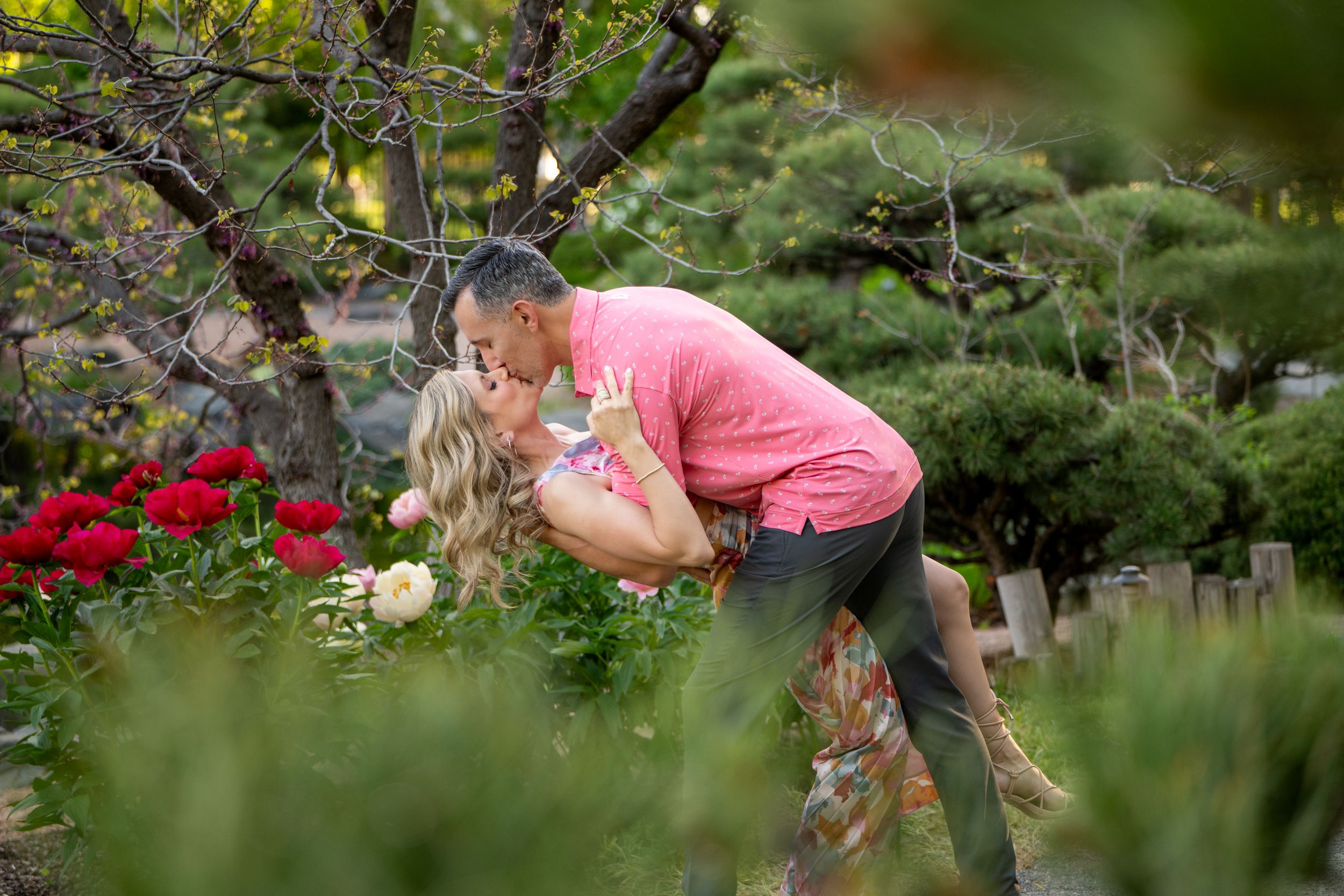Someone in a pink shirt stoops down to smell vibrant red flowers in a lush garden setting.