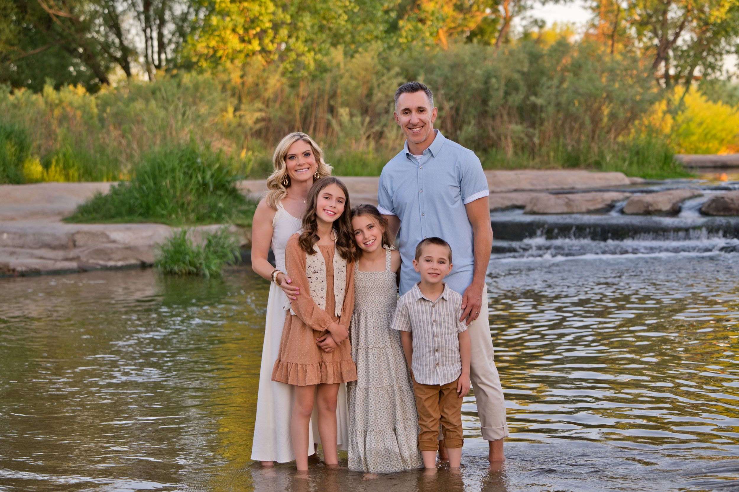 Family of five smiling together in a shallow river surrounded by lush greenery and a waterfall backdrop.
