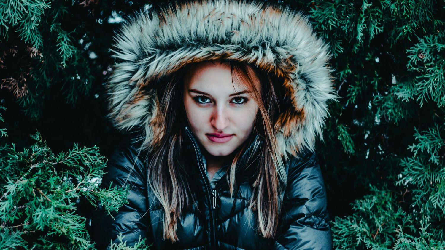 Winter portrait through evergreen branches with fur-trimmed hood in dark lighting.