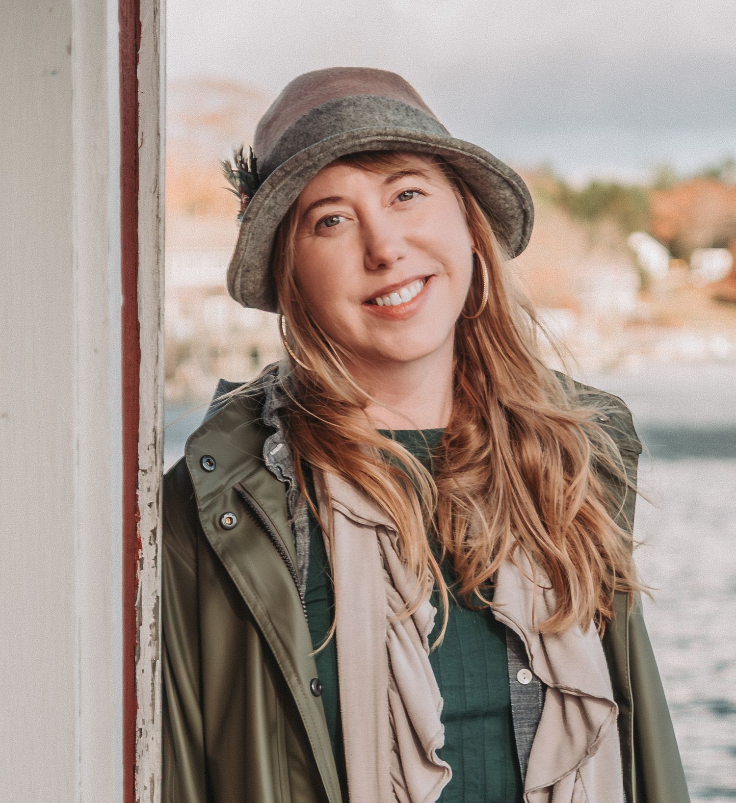 Person in olive green jacket and beige hat poses outdoors near waterfront on overcast day.