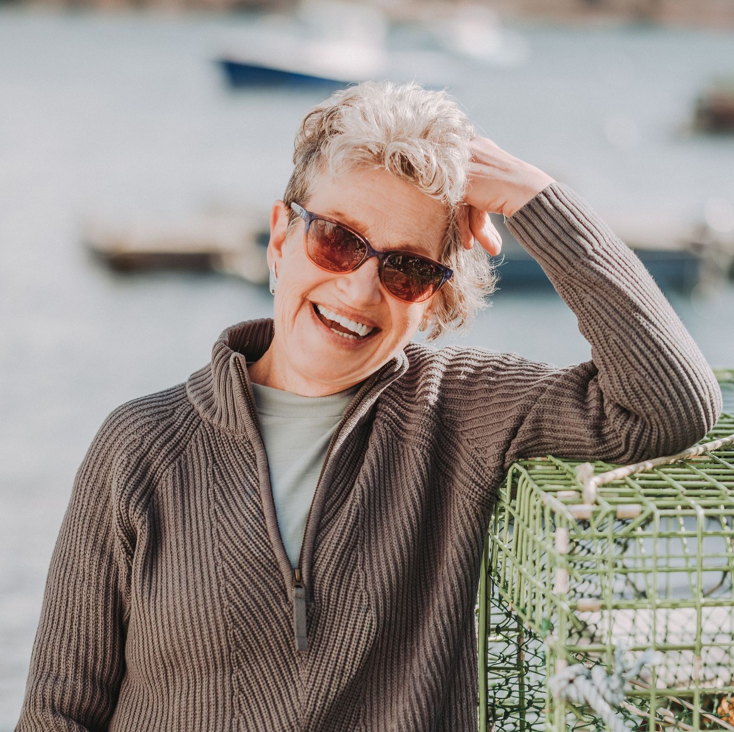 A veteran realtor in a brown sweater and sunglasses, laughing and enjoying a bright sunny day outdoors.