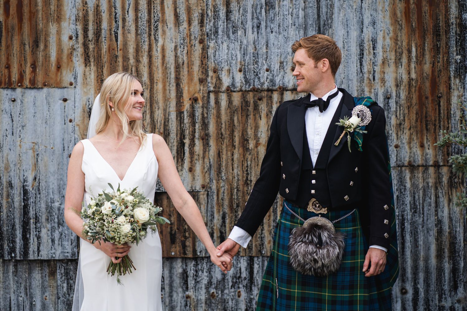 Bride and groom holding hands before rustic barn, groom wearing traditional Scottish tartan kilt, bride holding white bouquet.