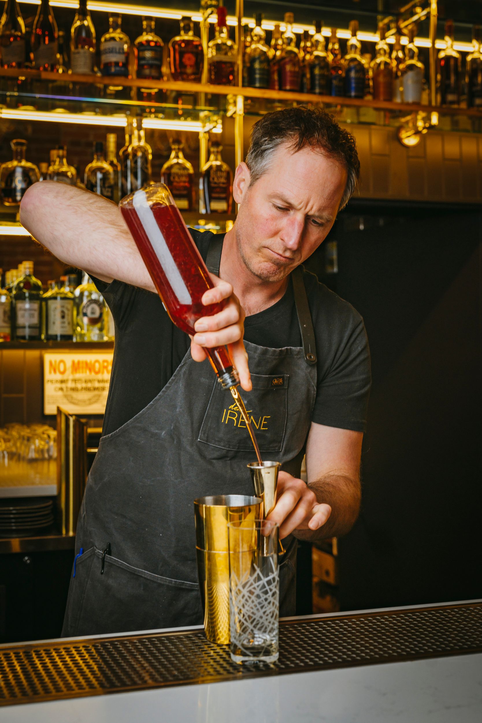 A bartender in black apron carefully pours a cocktail at a bar with illuminated shelves of liquor bottles in the background.