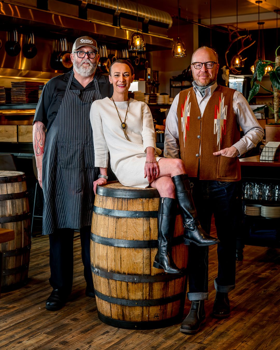 Three people in vintage-inspired attire pose together in a rustic bar setting with a wooden barrel and warm lighting.