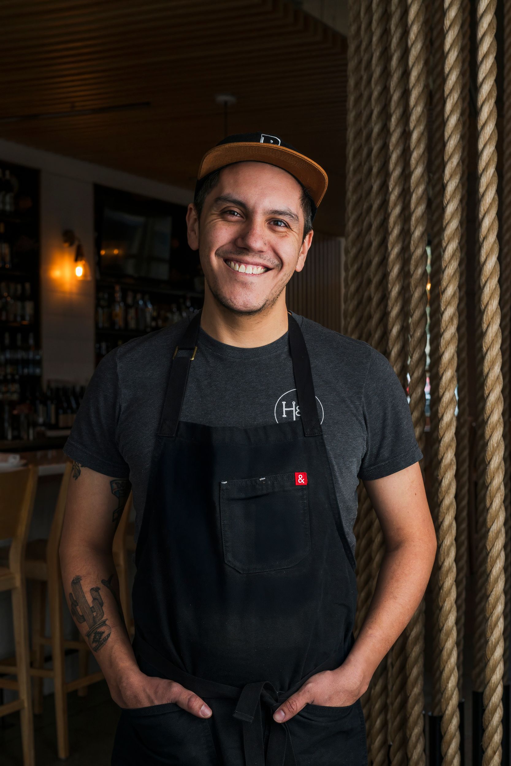 Restaurant worker in cap and black apron smiles while standing in dining area with rope decor and bar in background.