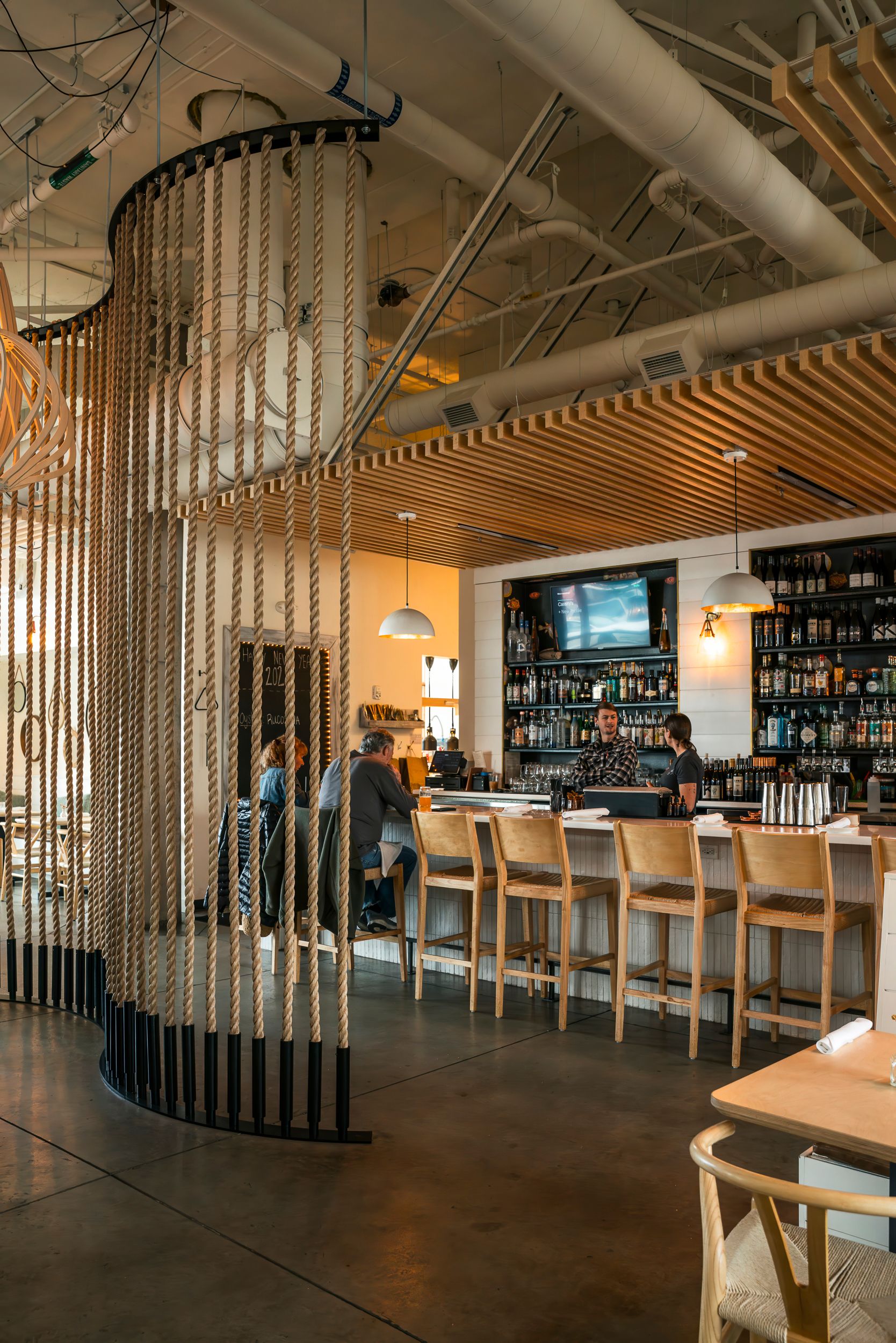 Modern restaurant bar interior with wooden slat ceiling, hanging chain divider, and fully stocked liquor shelves.