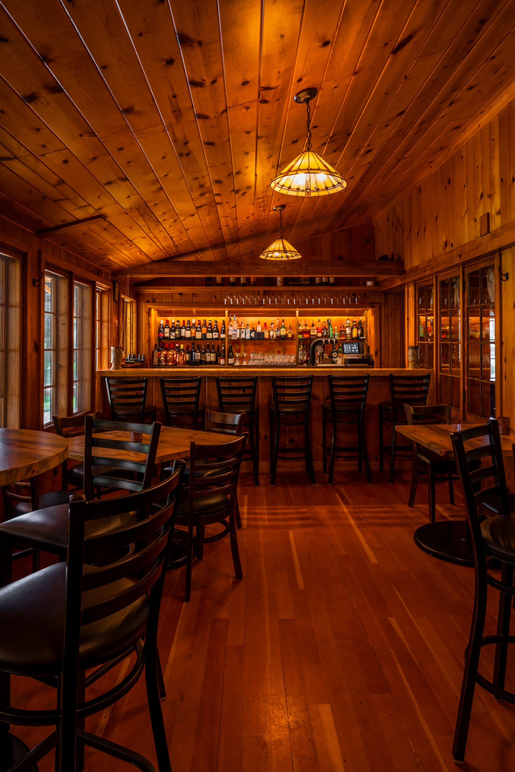 Cozy rustic bar interior with wooden walls, warm lighting from pendant lamps, and ladder-back chairs at a wooden counter.