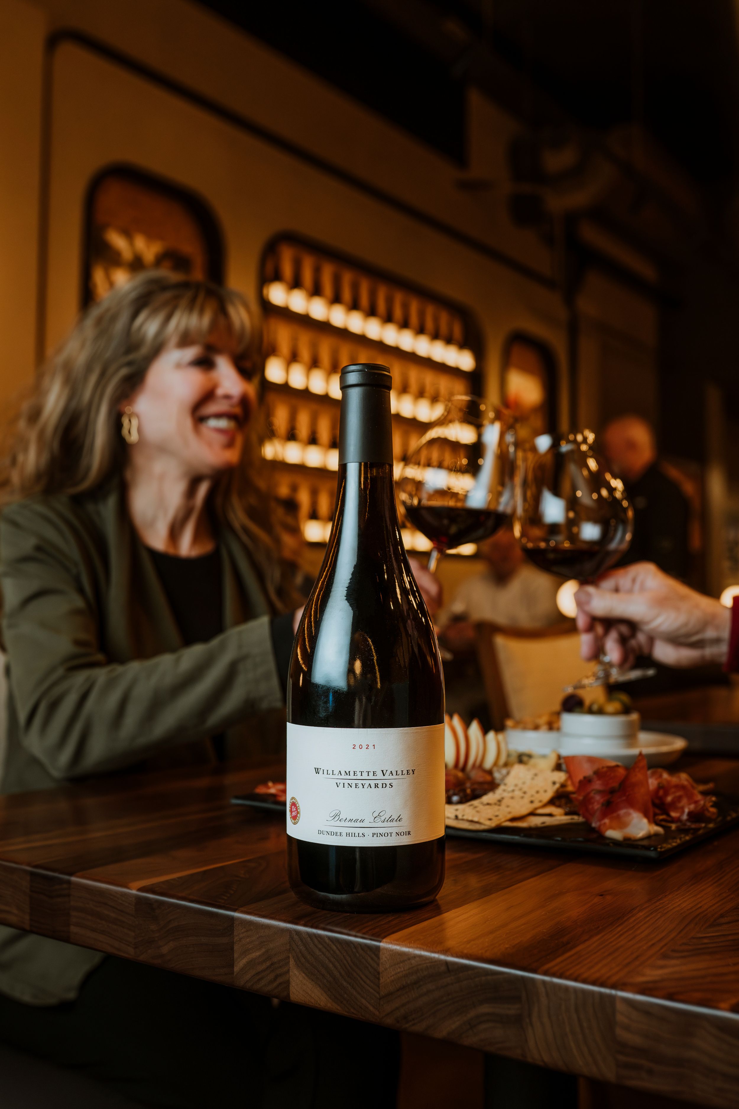 Wine enthusiasts gather at a cozy bar for a tasting, with a bottle on wooden counter and warm lighting in background.