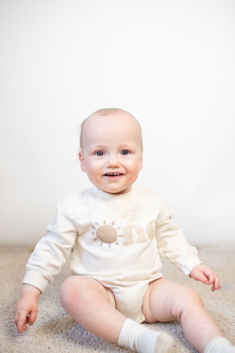 Baby wearing cream colored outfit sitting on carpet in progressive poses.