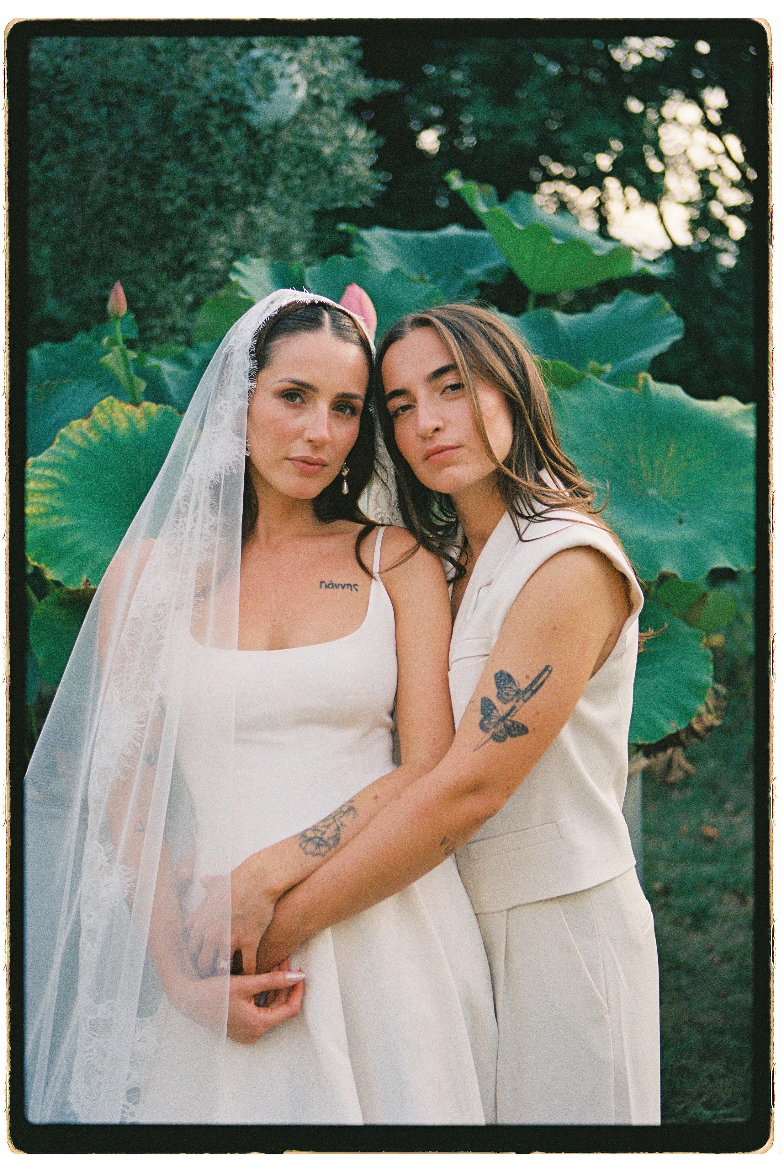 Two brides in white wedding attire pose together outdoors near large tropical leaves.