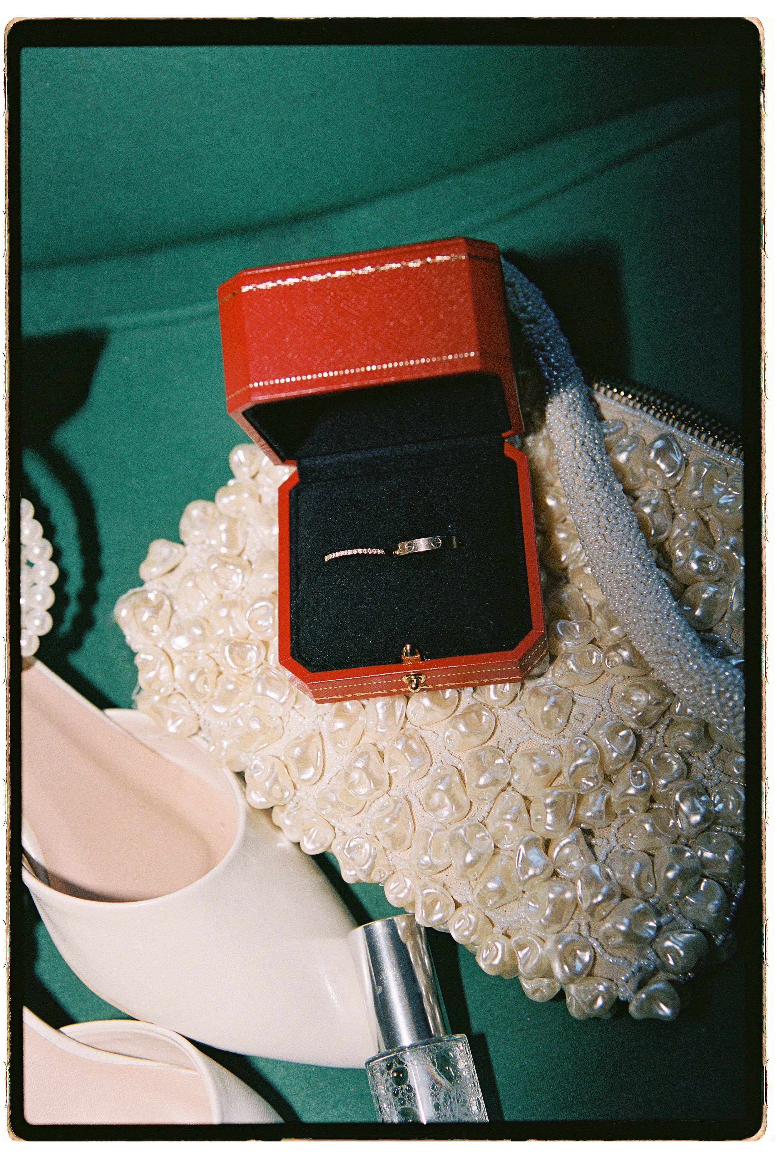 A silver ring in a red Cartier box displayed on a pearl-embellished accessory with white heels on green background.