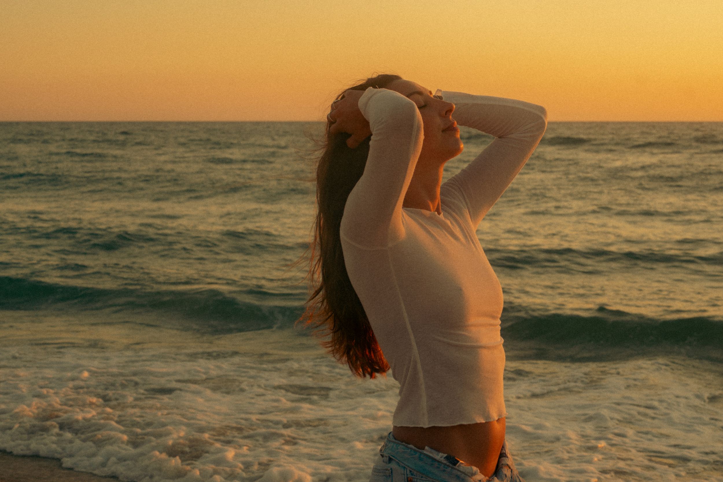Woman with long hair poses on beach at golden sunset, wearing white top and denim jeans with ocean waves behind her.