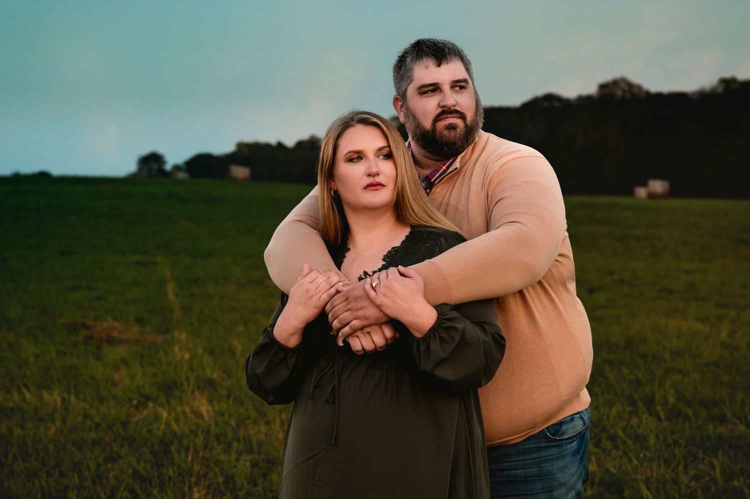 Couple embracing in green field at dusk, man holding woman from behind in romantic outdoor portrait.
