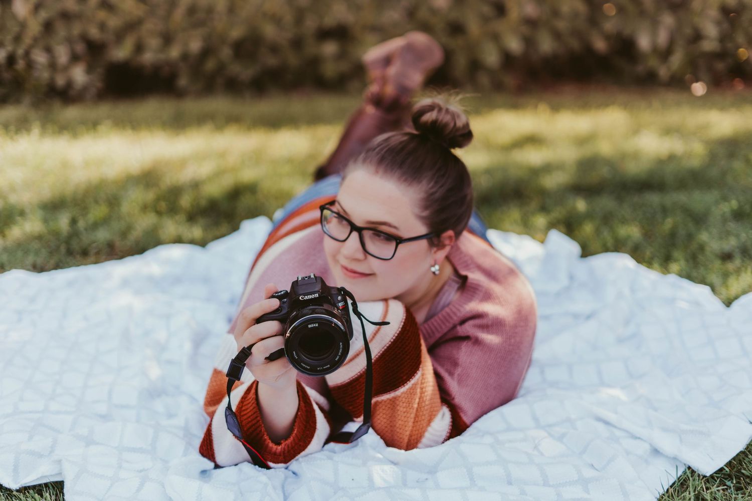 Young woman with glasses lying on blanket outdoors holding a Canon DSLR camera.