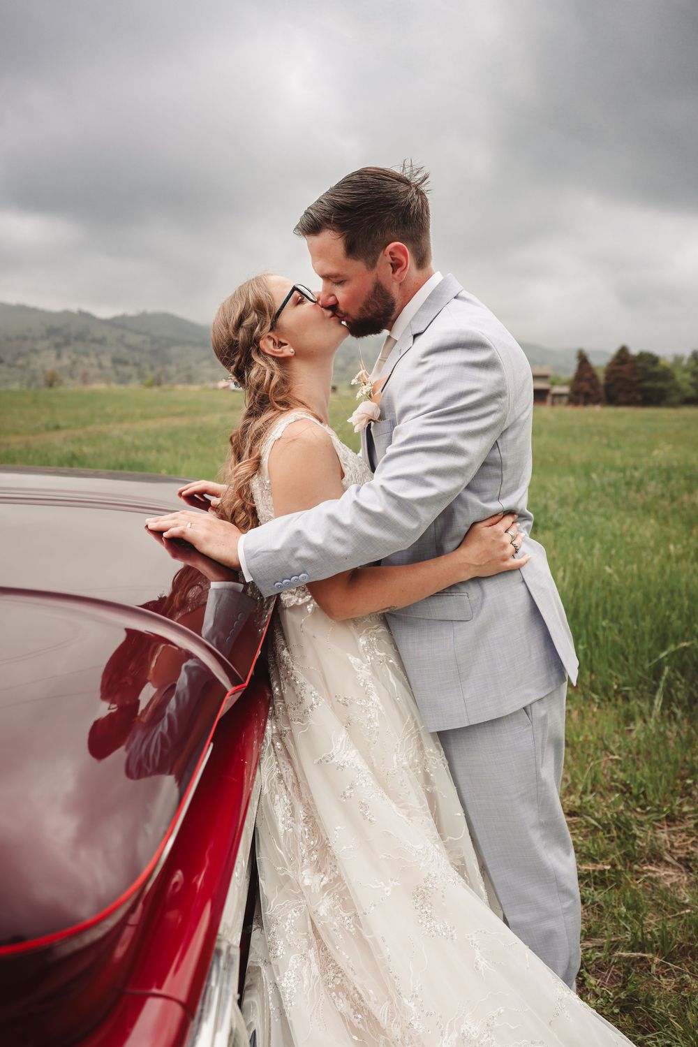 Newlywed couple sharing an intimate moment beside a vintage red car in a rustic countryside setting.