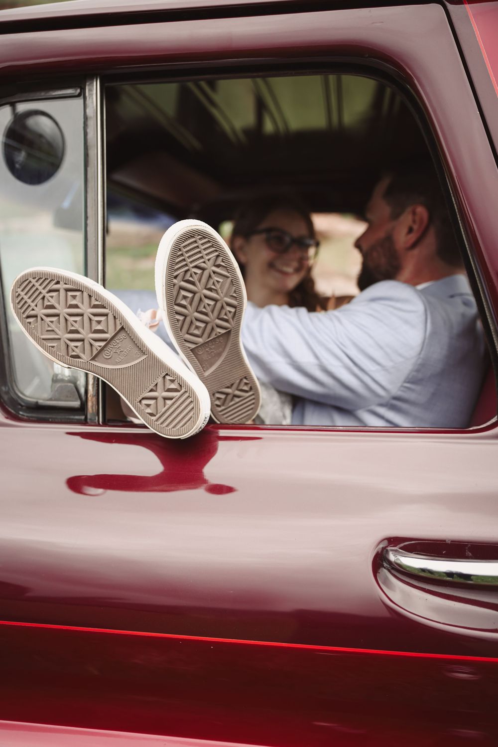 The soles of white sneakers rest on a red vintage car window while people are seated inside.
