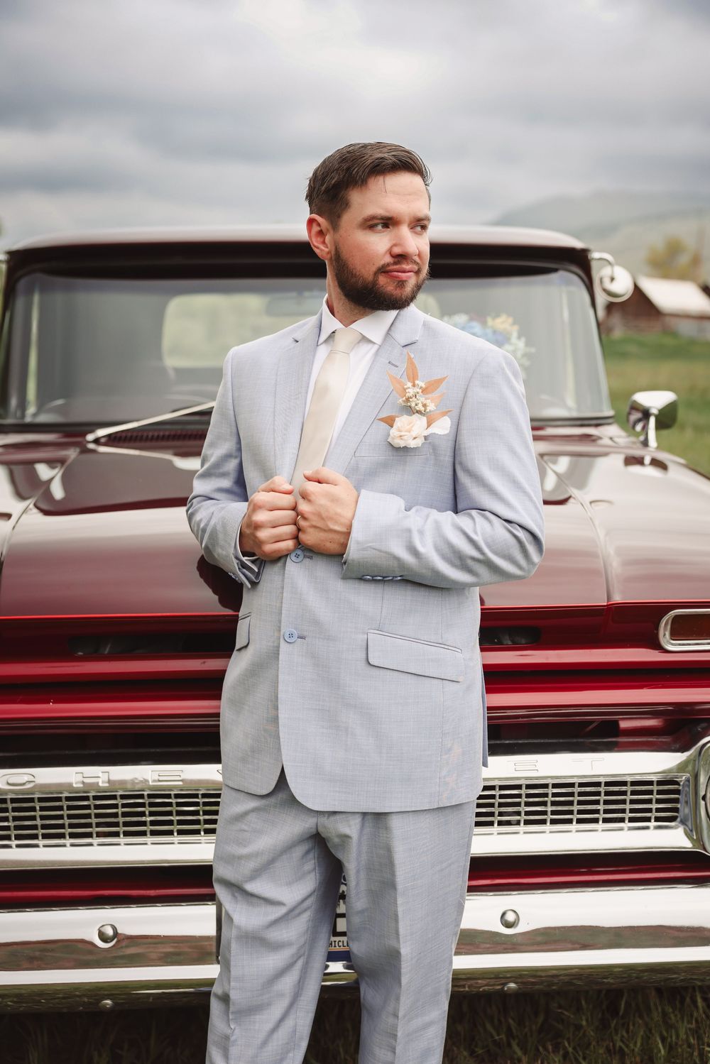 A groom in a light gray suit and blush boutonniere stands in front of a vintage maroon truck at an outdoor wedding venue.