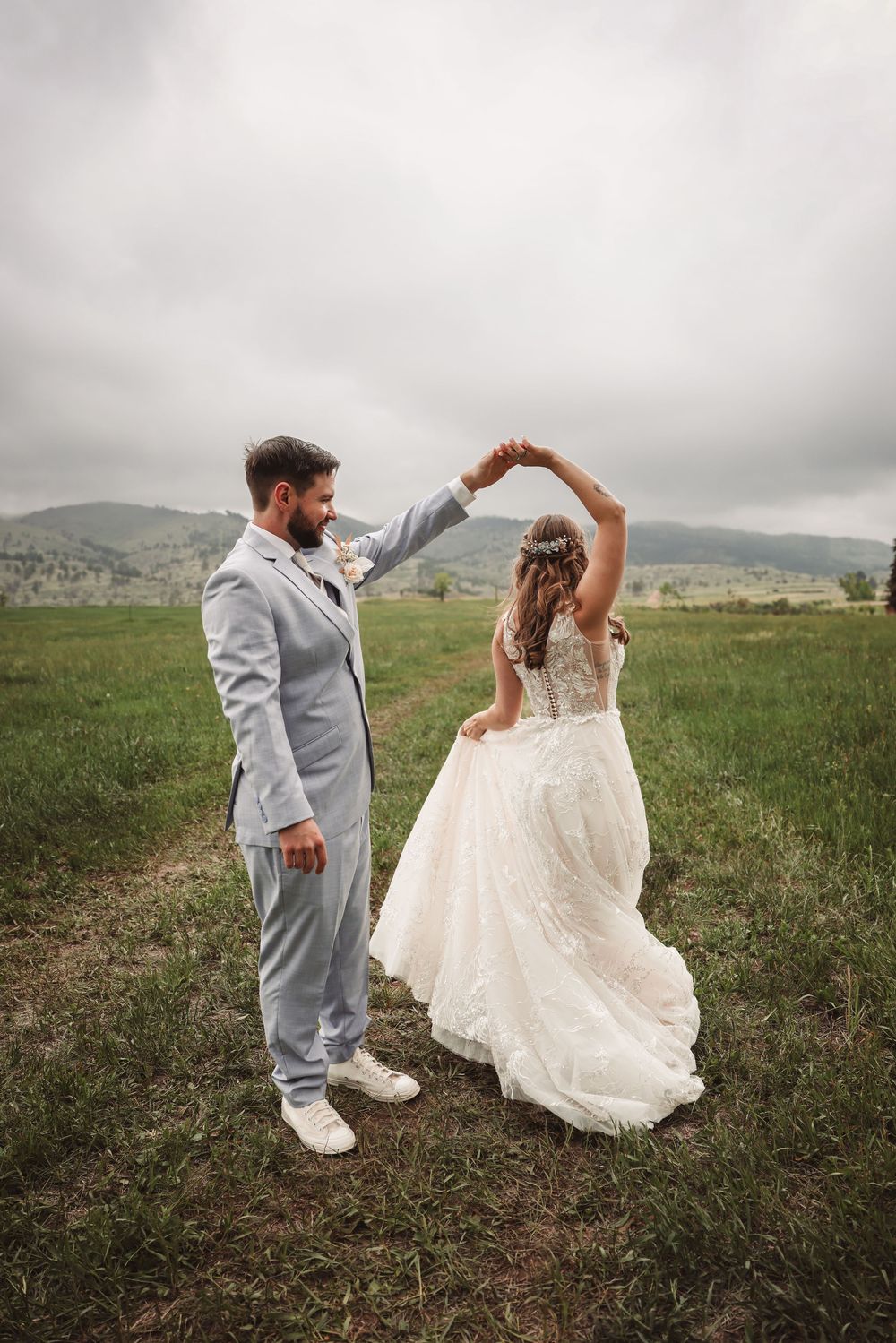 A couple in wedding attire dance together in a meadow with mountains in the background on an overcast day.