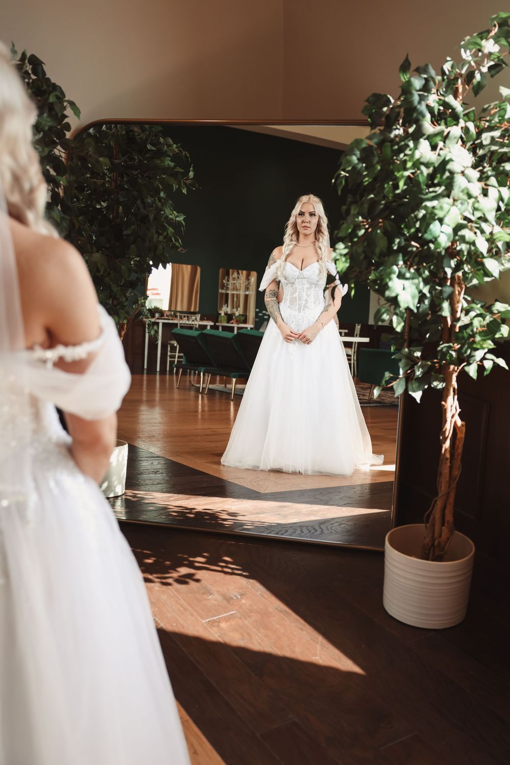 A white wedding dress reflects in a mirror with decorative potted trees and wooden flooring in an elegant indoor setting.