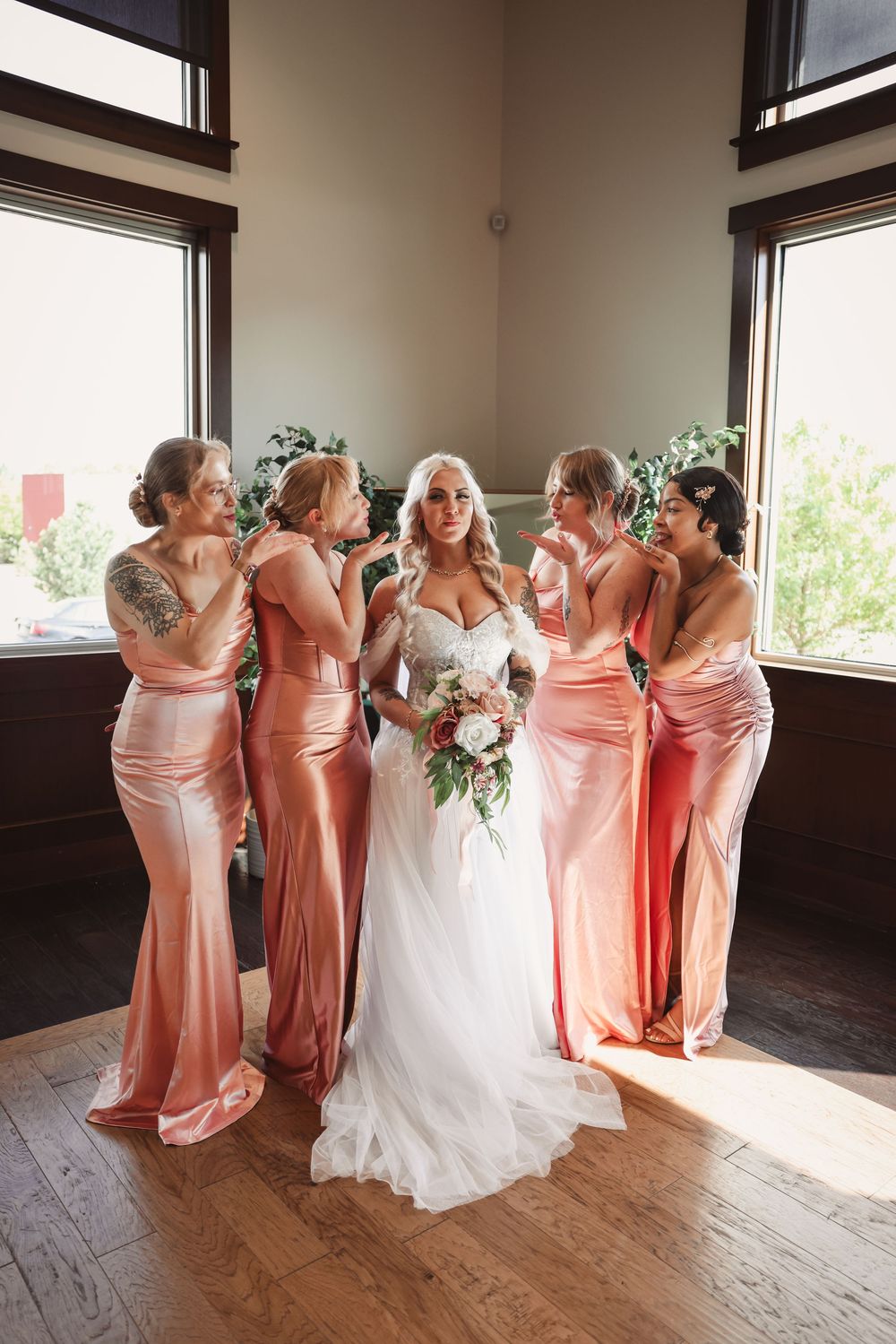 A wedding party in peach-colored bridesmaid dresses stands together indoors with natural light streaming through windows.