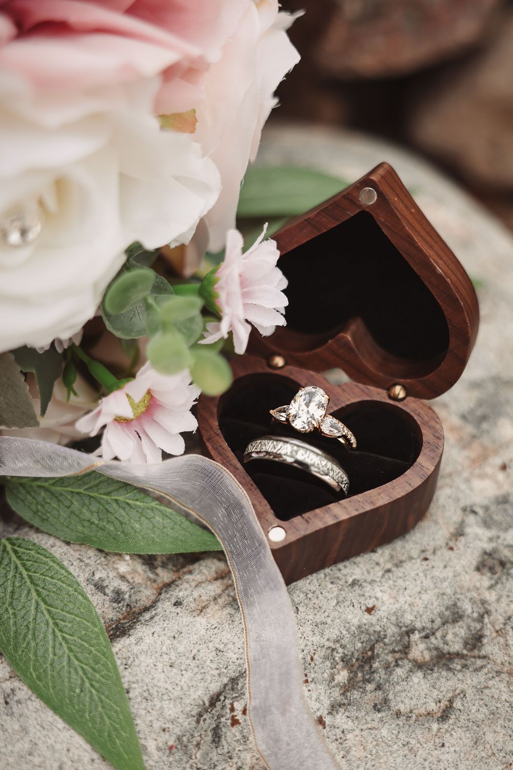 Two diamond engagement rings nestled in a wooden ring box with delicate pink flowers and green leaves.