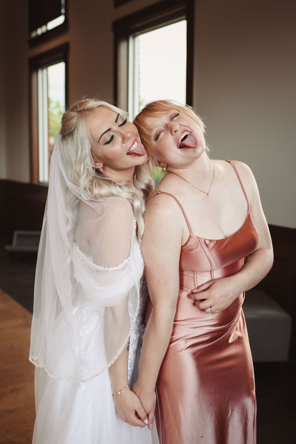 Two women in matching light-colored dresses share a joyful laugh together at an indoor celebration.
