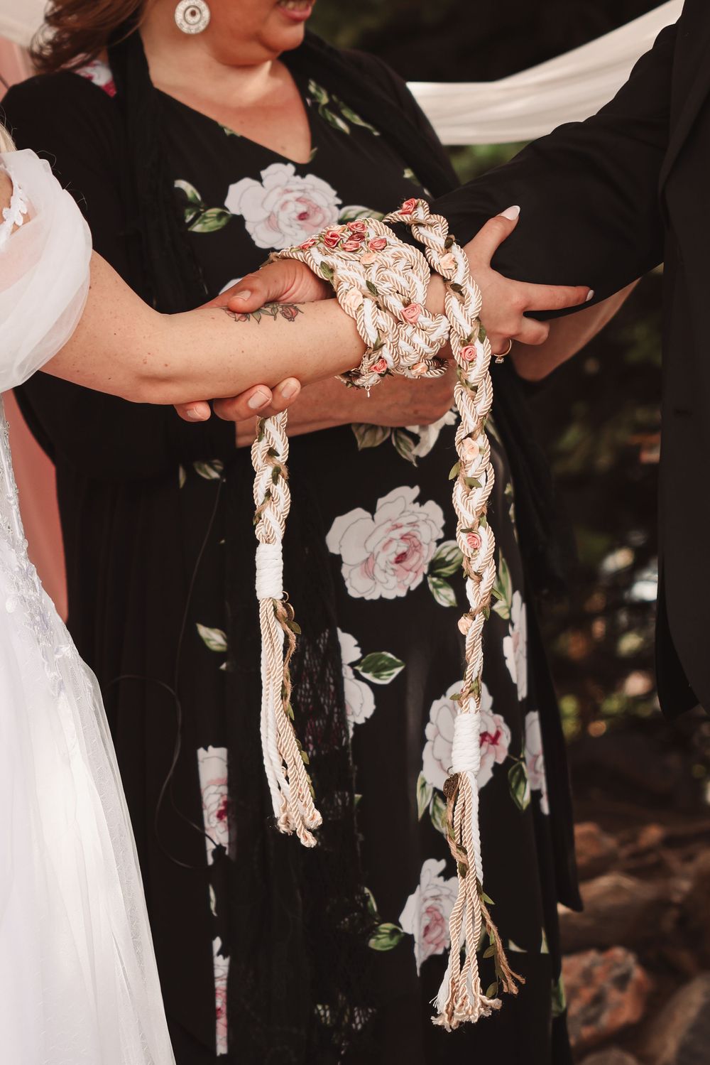 Hands holding a decorative braided rope or cord with tassels against a black floral dress at a wedding ceremony.