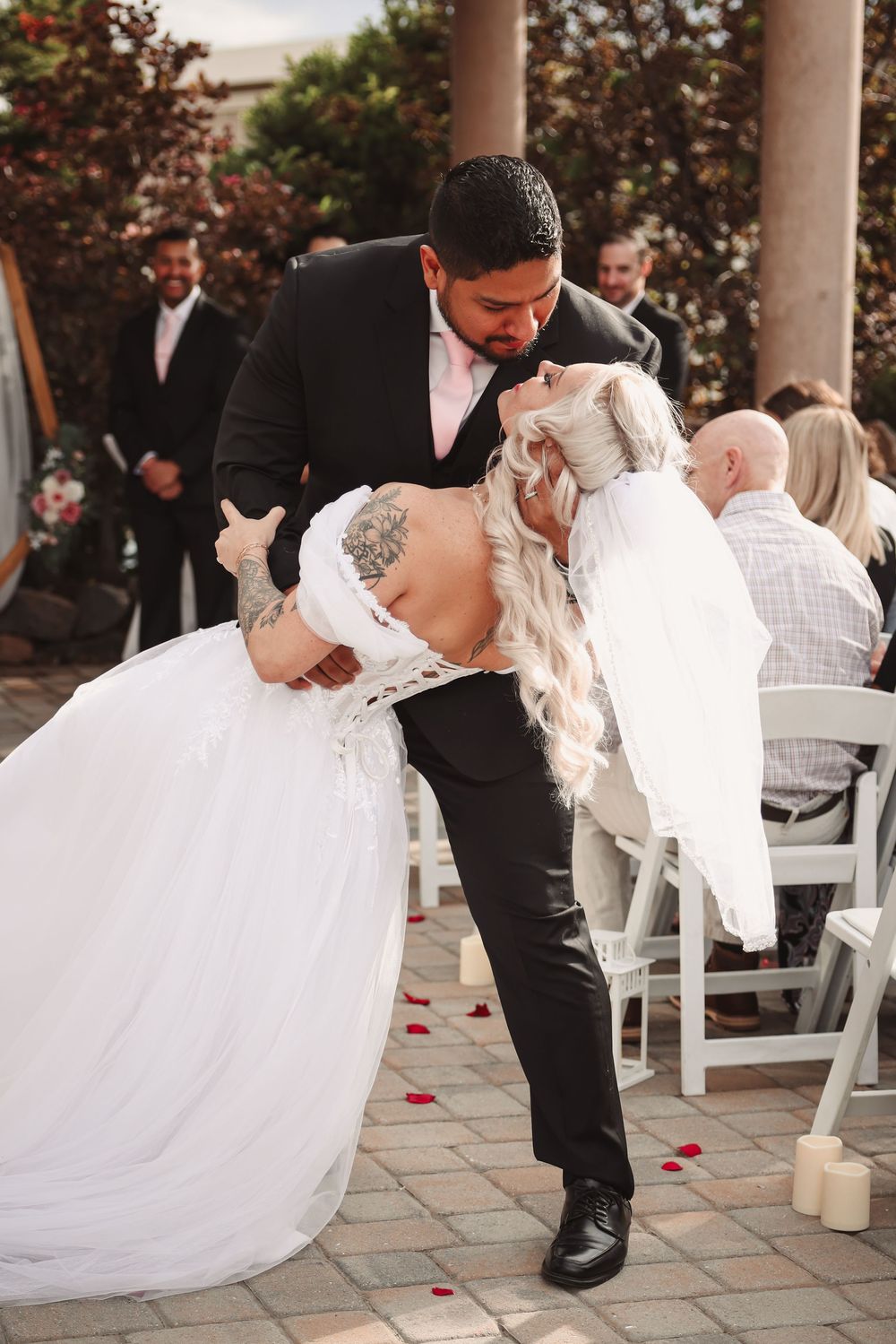 Couple shares romantic dip kiss at outdoor wedding ceremony with red rose petals scattered on brick patio.