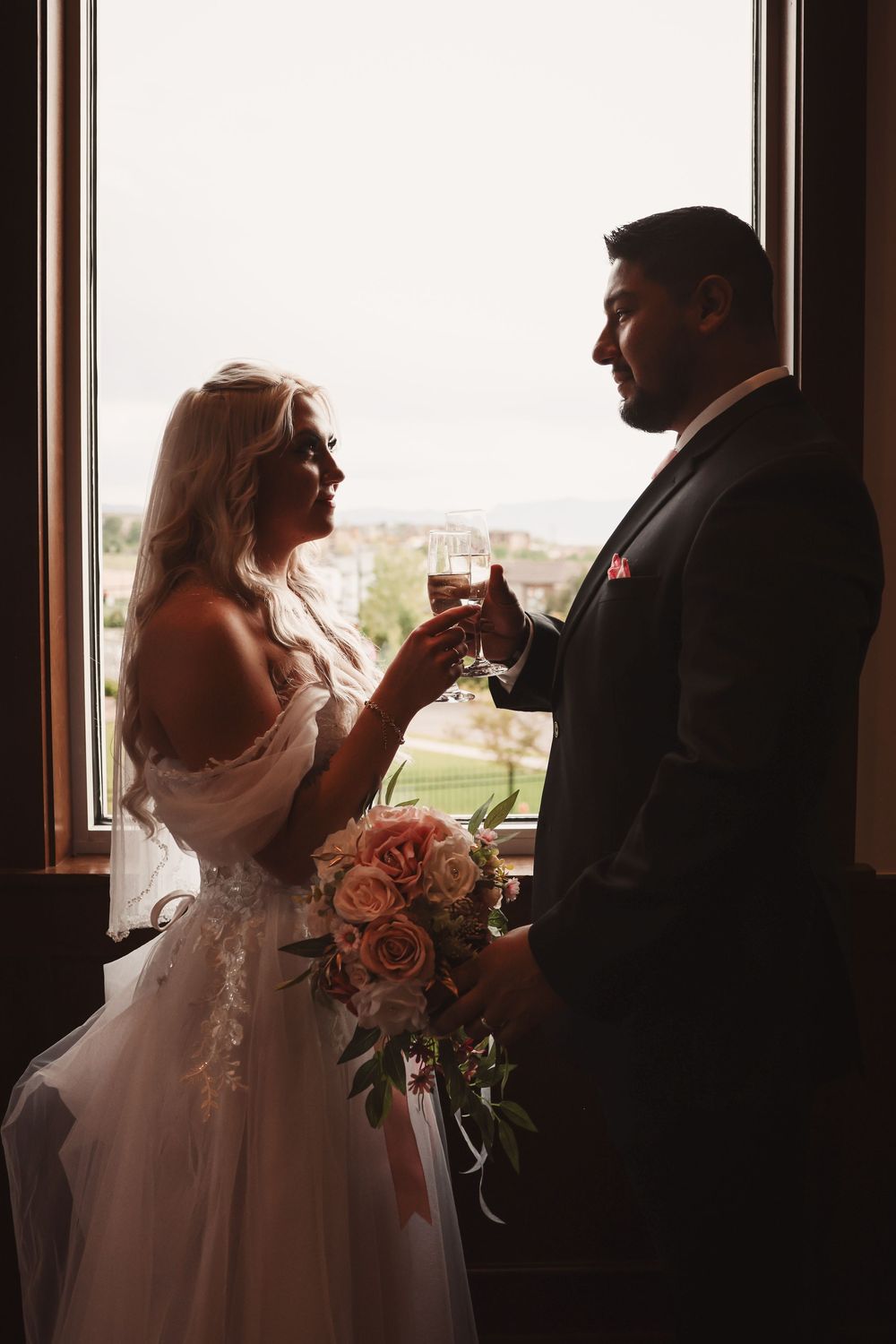 Silhouetted couple sharing champagne toast by window as sunlight streams in, with pink wedding bouquet visible.