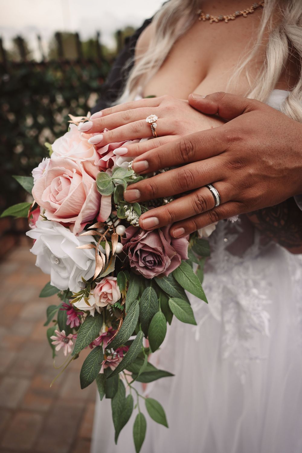 Close-up of hands with engagement ring holding pink and mauve roses with greenery against white wedding dress.