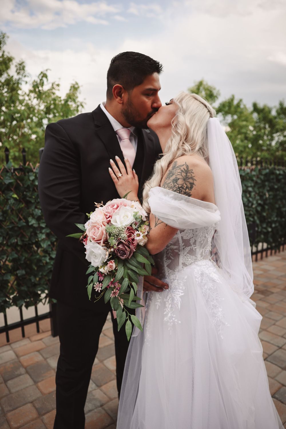 Bride and groom share romantic kiss outdoors with a pink floral bouquet and garden backdrop.