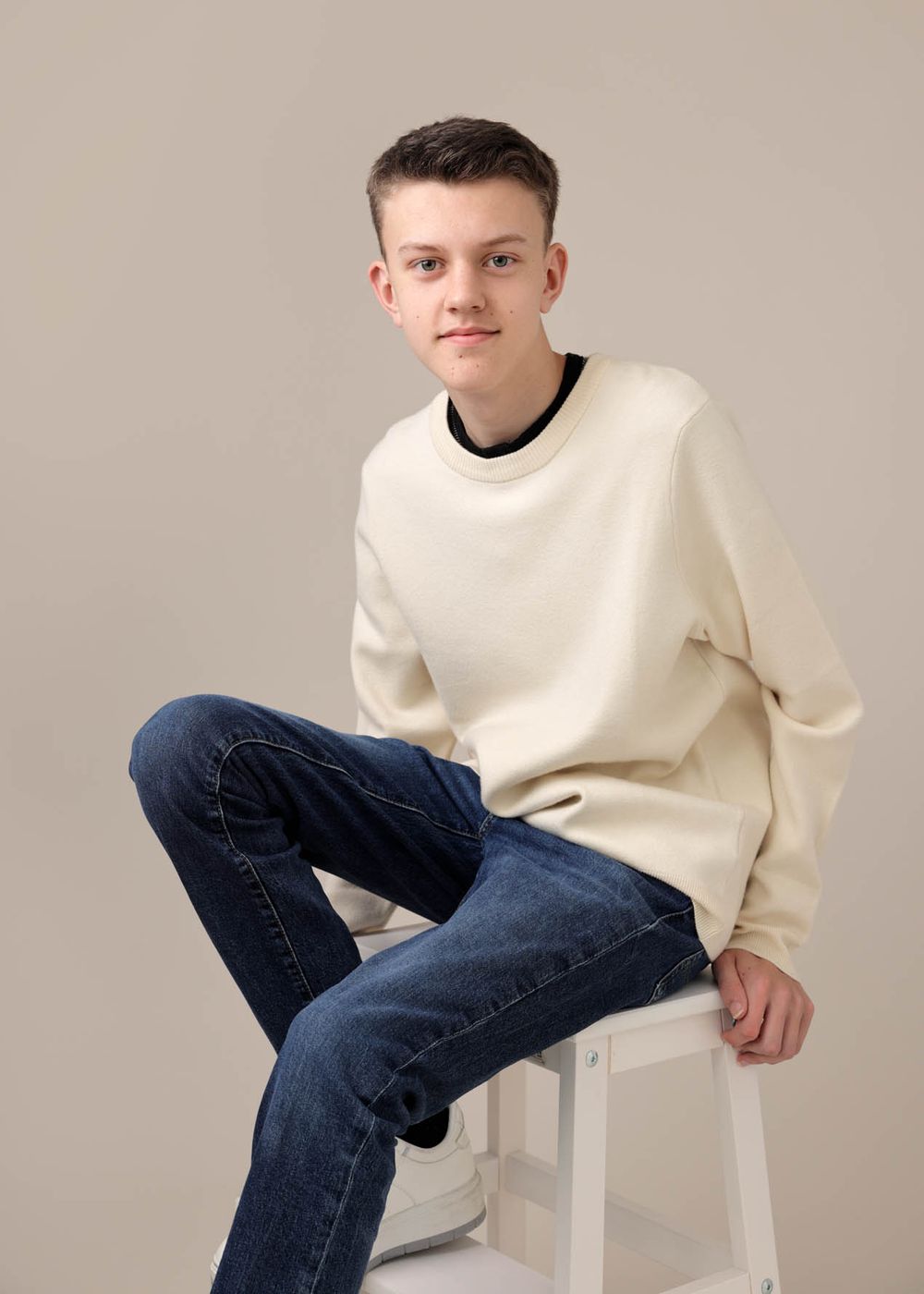 Teen boy in cream sweater and blue jeans sits on white stool, smiling at camera in studio model shoot