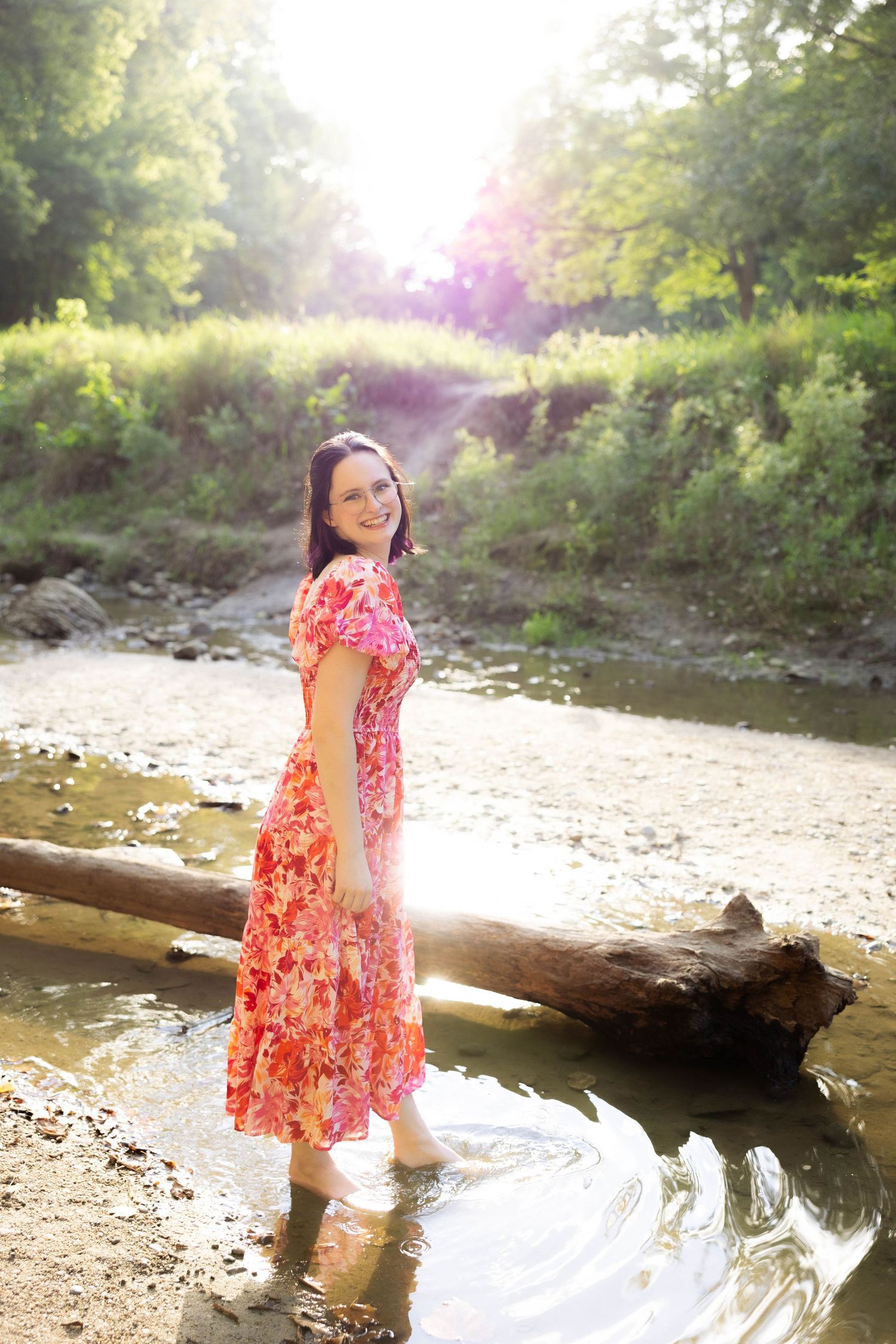 Woman in floral dress stands by creek in sunlit forest setting.