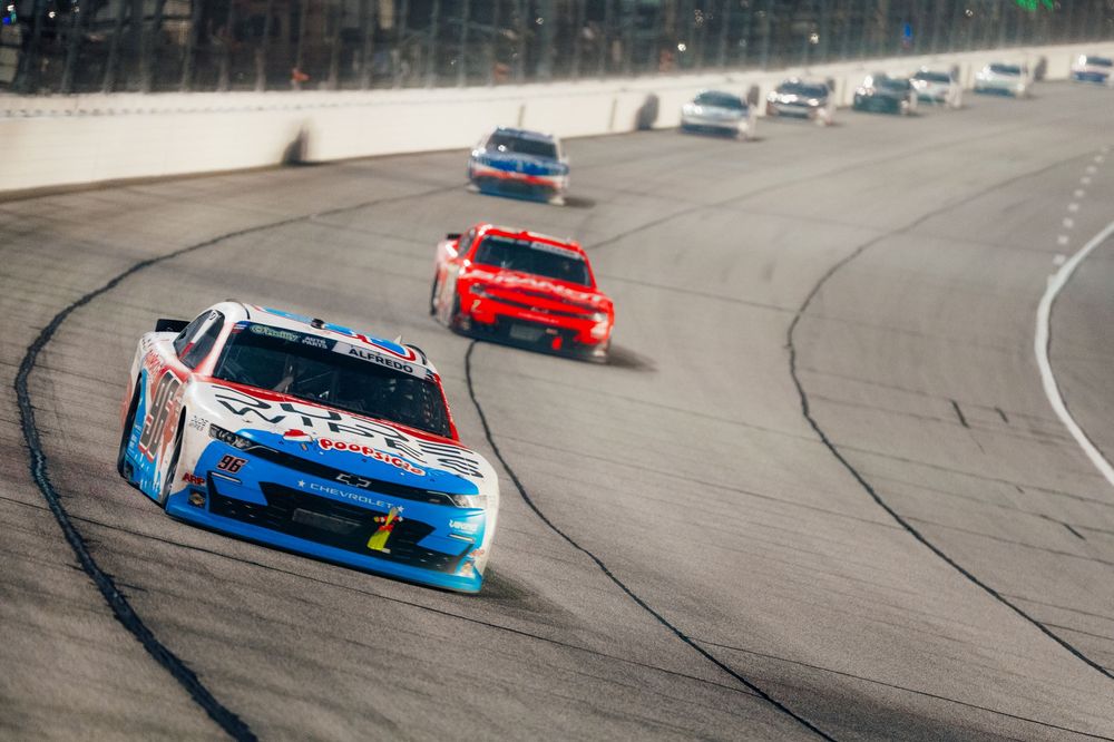 NASCAR race cars speed around a banked oval track under bright lights during an exciting nighttime stock car racing event.