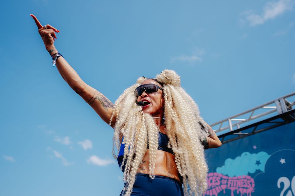 Energetic performer with blonde braids raises hand at outdoor music festival under blue sky.