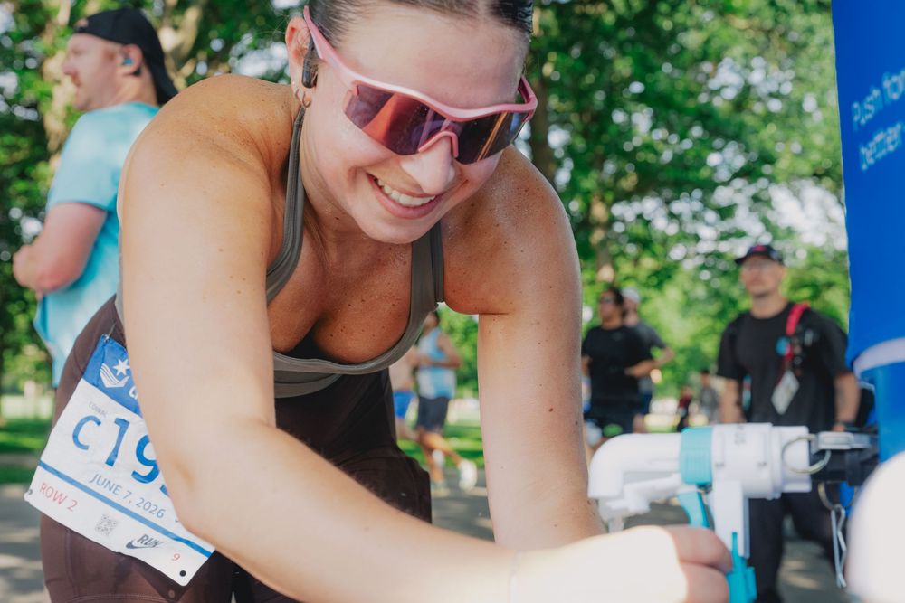 Smiling woman with pink sunglasses wearing race bib C19 leaning over at an outdoor running event.