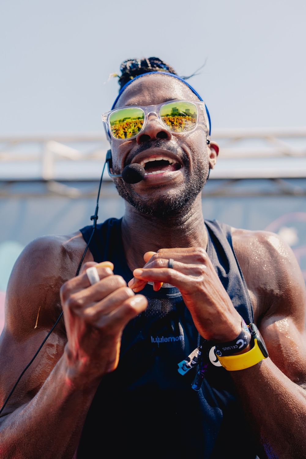 Athletic man with yellow sunglasses and yellow watch smiling during outdoor sports event, wearing earphones and vest.