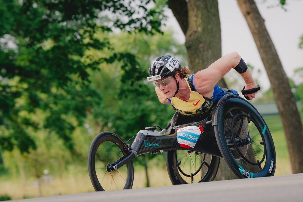 Athlete competing in a wheelchair race on a road, wearing a helmet and racing gloves, surrounded by green trees.
